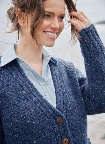 A woman with brown hair wearing a blue wool cardigan is standing on a beach with the ocean behind her.
